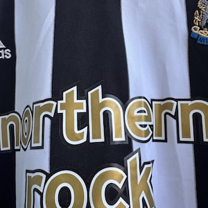 Może przedstawiać: Czarno-biała koszulka piłkarska w paski ze złotym napisem "northern ROCK". Koszulka posiada logo drużyny i logo Adidas.