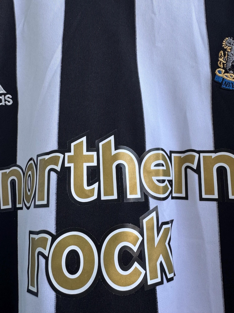 Może przedstawiać: Czarno-biała koszulka piłkarska w paski ze złotym napisem "northern ROCK". Koszulka posiada logo drużyny i logo Adidas.