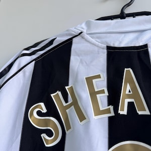 Może przedstawiać: Czarno-biała koszulka piłkarska w paski z napisem "SHEA" złotymi literami. Koszulka wisi na czarnym wieszaku na białej ścianie. Logo Adidas jest widoczne na ramieniu.