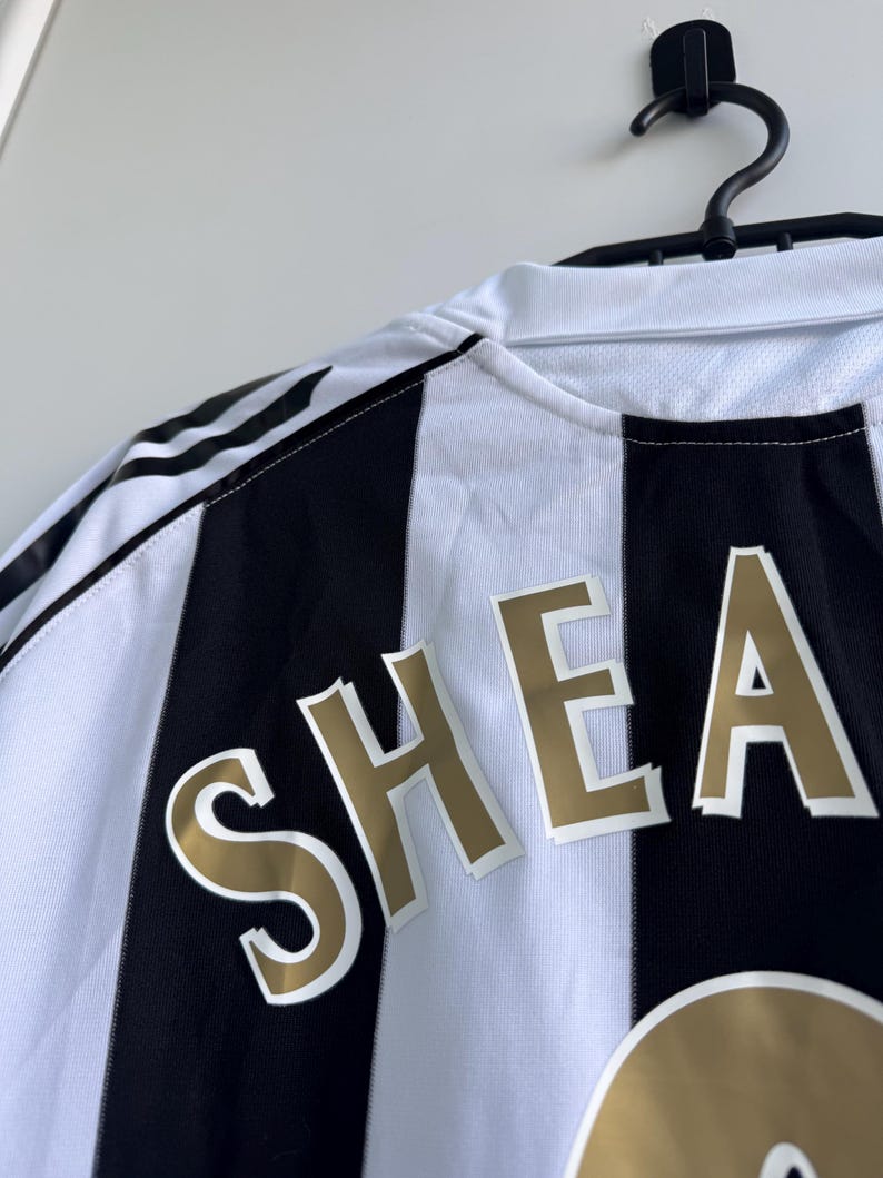 Może przedstawiać: Czarno-biała koszulka piłkarska w paski z napisem "SHEA" złotymi literami. Koszulka wisi na czarnym wieszaku na białej ścianie. Logo Adidas jest widoczne na ramieniu.