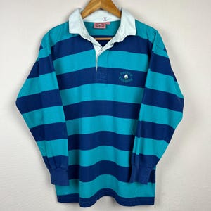 Puede incluir: Una camiseta de rugby de manga larga con rayas horizontales en verde azulado y azul marino. La camiseta tiene un cuello blanco y un pequeño logotipo bordado. El texto "COTTON TRADERS" es visible en la camiseta.