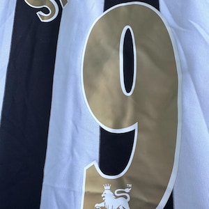 Może przedstawiać: Zbliżenie na czarno-białą koszulkę piłkarską w paski. Numer 9 jest złoty, z napisem "SHEARER" u g&oacute;ry. Logo Premier League znajduje się na dole.