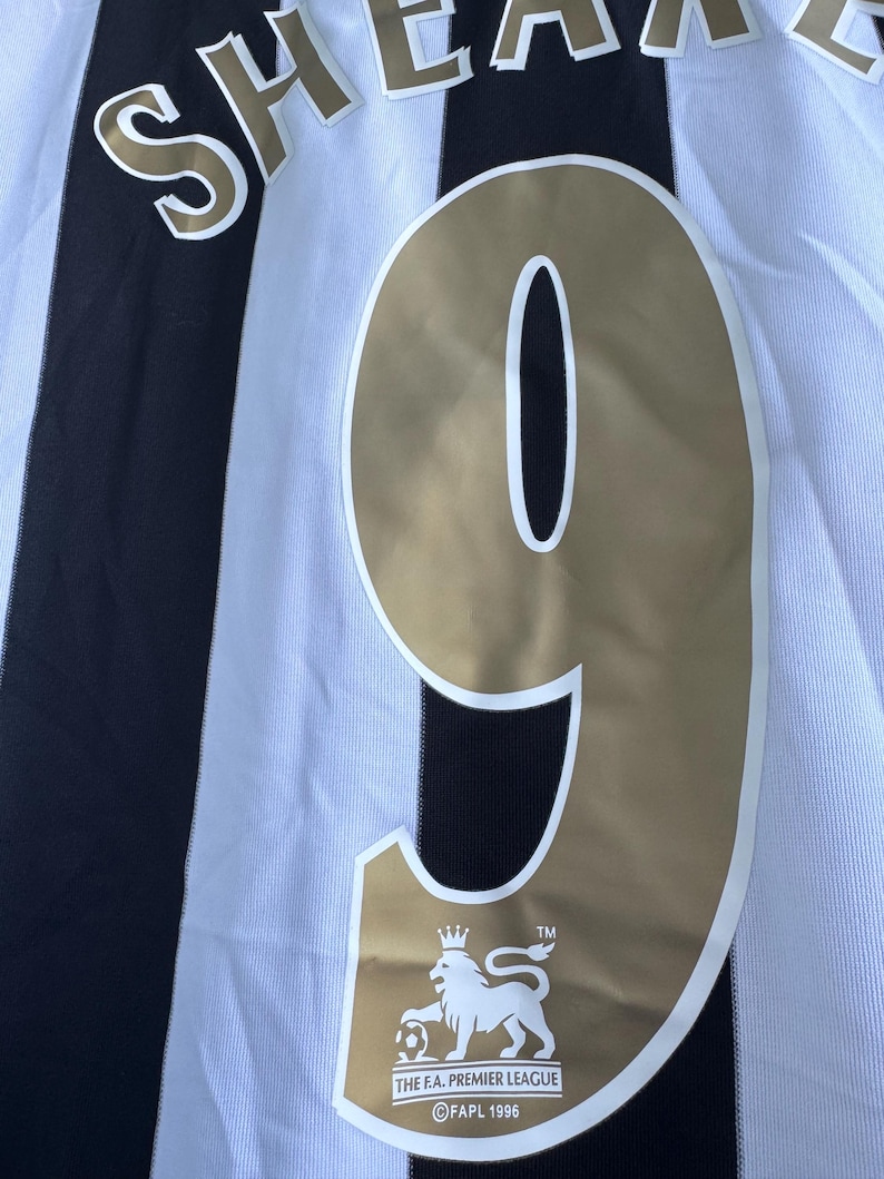 Może przedstawiać: Zbliżenie na czarno-białą koszulkę piłkarską w paski. Numer 9 jest złoty, z napisem "SHEARER" u g&oacute;ry. Logo Premier League znajduje się na dole.
