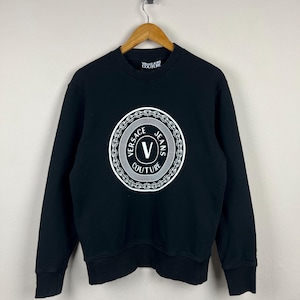 Puede incluir: Una taza de cerámica blanca con asa negra y el texto "But First, Coffee" en negrita negra. La taza se encuentra sobre un fondo borroso, sugiriendo un ambiente acogedor.