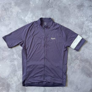 May include: A short-sleeve cycling jersey in a muted purple color with a full-length front zipper. The garment features a white horizontal band on the left sleeve and the brand name Rapha printed in white on the upper left chest area.