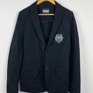 Puede incluir: Blazer negro con cierre de dos botones, solapas con muescas y dos bolsillos delanteros. Presenta un escudo en el pecho y una etiqueta en el cuello. El blazer está colgado en una percha de madera.