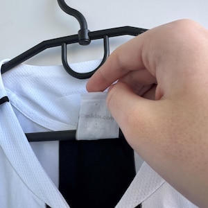 Może przedstawiać: Biało-czarna koszulka sportowa wisząca na czarnym wieszaku. Koszulka ma czarny pasek i biały kołnierz. Etykieta zawiera napis adidas.com.