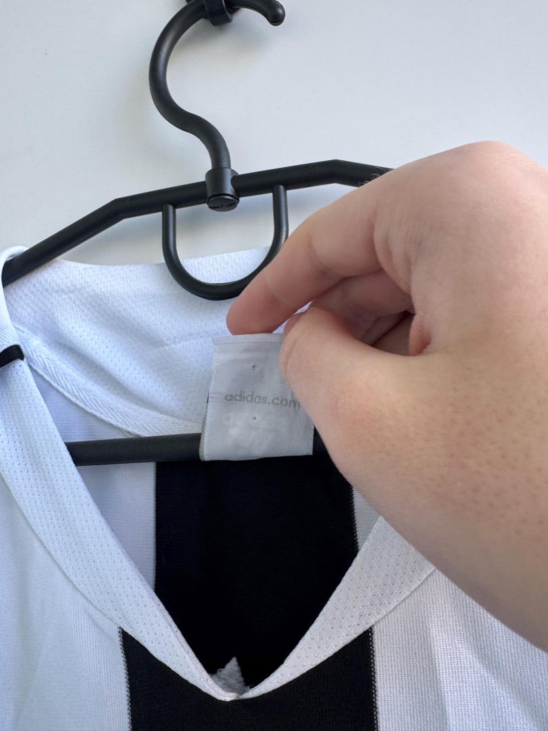 Może przedstawiać: Biało-czarna koszulka sportowa wisząca na czarnym wieszaku. Koszulka ma czarny pasek i biały kołnierz. Etykieta zawiera napis adidas.com.