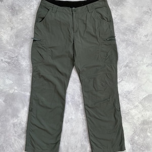 May include: Olive green cargo pants featuring a button and zipper fly closure, belt loops, two side pockets, and two zippered cargo pockets on the thighs. The pants have a black interior waistband lining and straight legs with reinforced knee panels.