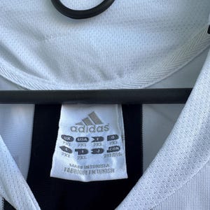 Może przedstawiać: Zbliżenie białej i czarnej koszulki piłkarskiej Adidas. Koszulka ma biały kołnierz i czarno-biały wz&oacute;r w paski. Widoczne jest logo Adidas i metka z rozmiarem.