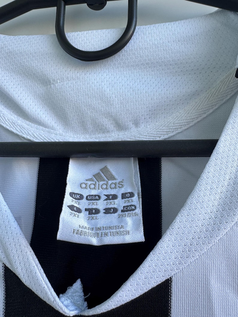 Może przedstawiać: Zbliżenie białej i czarnej koszulki piłkarskiej Adidas. Koszulka ma biały kołnierz i czarno-biały wz&oacute;r w paski. Widoczne jest logo Adidas i metka z rozmiarem.