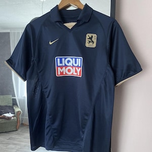 May include: Navy blue athletic jersey with gold trim and a collared neckline. Features the Nike logo, a team crest, and the "LIQUI MOLY" logo. The jersey is hanging on a wooden hanger.
