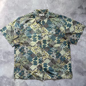 Può includere: Una camicia a maniche corte con bottoni e un motivo patchwork multicolore nei toni del verde acqua, verde oliva e crema. Il design include vari motivi floreali, geometrici e astratti disposti a griglia. Un'etichetta Guess si trova sul colletto.