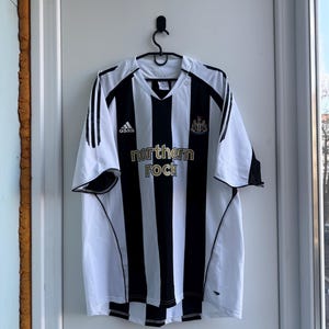 Może przedstawiać: Czarno-biała koszulka piłkarska w paski z napisem "northern rock" w kolorze złotym. Koszulka ma dekolt w serek i logo Adidas. Herb Newcastle United znajduje się na prawej piersi.
