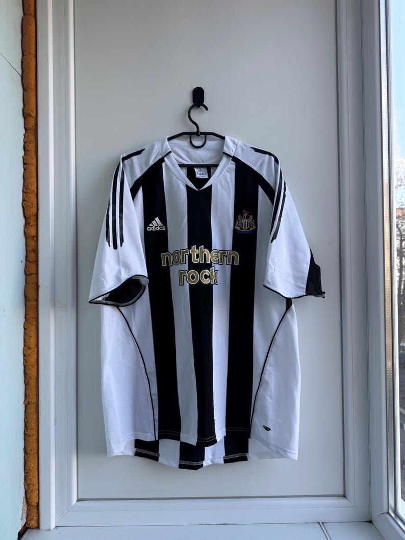 Może przedstawiać: Czarno-biała koszulka piłkarska w paski z napisem "northern rock" w kolorze złotym. Koszulka ma dekolt w serek i logo Adidas. Herb Newcastle United znajduje się na prawej piersi.