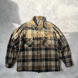 Pode incluir: Uma camisa de manga comprida com botões e um padrão xadrez em tons de castanho, bege e preto. A camisa inclui um colarinho padrão, dois bolsos no peito com abas abotoadas e um fecho frontal de botões em toda a sua extensão.