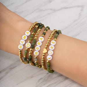 May include: Several beaded bracelets with gold accents and green beads are displayed. Some bracelets spell out "MAMA", "NORA", and "LUKE" with white letter beads. A gold heart charm is also visible.