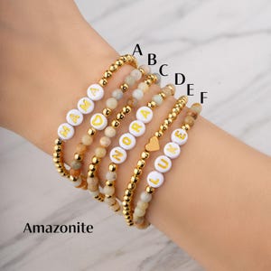 May include: A collection of beaded bracelets with gold accents and white letter beads spelling out names. The bracelets feature a mix of gold beads and natural stone beads. The word "Amazonite" is printed at the bottom of the image.