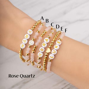 May include: Six beaded bracelets with white letter beads spelling out "MAMA", "NORA", and "LUKE", and a heart charm. The bracelets are gold and rose quartz. The text "Rose Quartz" is at the bottom.