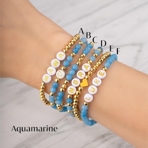 May include: Six beaded bracelets with the word "MAMA", "NORA", and "LUKE" spelled out in white letter beads. The bracelets are made of gold-colored beads and aquamarine-colored beads. The word "Aquamarine" is written at the bottom of the image.