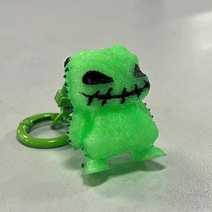 May include: A neon green figurine of a character with black stitched details and a stitched mouth. The figurine is attached to a green metal keyring. The character has a textured surface and is standing on a light gray surface.