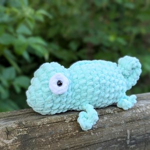 May include: A light blue crochet chameleon toy with a white eye and black pupil. The plush reptile has a textured, fuzzy appearance and is lying on a wooden surface. The toy is a soft sculpture, suitable for children or as a decorative item.