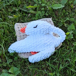 May include: A handmade white plush goose toy with an orange beak and black eyes, resting on a beige crocheted square. The soft, fluffy goose is lying on a bed of green grass.