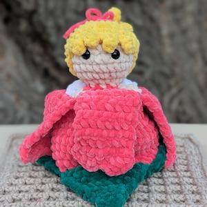 May include: A handmade crochet doll with a yellow head, pink bow, and a pink flower-shaped dress. The doll is resting on a beige crocheted square with a green leaf-shaped base.