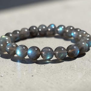 May include: A stretch bracelet composed of round, polished labradorite beads. The beads are primarily dark grey with iridescent flashes of blue and green light reflecting off their surfaces, creating a shimmering effect across the circular band.