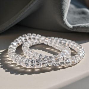 May include: Two clear, spiral hair ties with a square, faceted design. The ties are transparent and reflect light, creating a sparkling effect. The background includes a soft, grey fabric and a neutral surface.