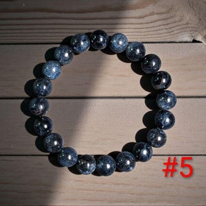 Puede incluir: Una pulsera circular compuesta por cuentas esf&eacute;ricas pulidas en tonos azul oscuro, gris y negro con una textura jaspeada. Las cuentas est&aacute;n ensartadas en un bucle continuo. El texto rojo #5 es visible en la esquina inferior derecha.