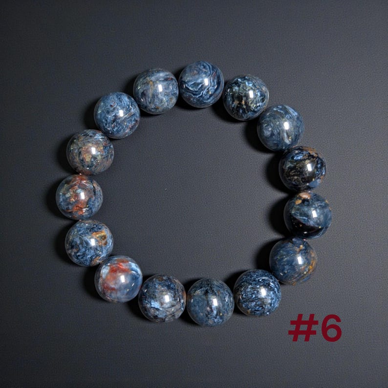 Puede incluir: Una pulsera circular compuesta por cuentas esf&eacute;ricas pulidas de piedra pietersita azul. Las cuentas presentan patrones arremolinados de azul profundo, gris y vetas ocasionales de color marr&oacute;n rojizo. El texto #6 es visible en la esquina inferior derecha.
