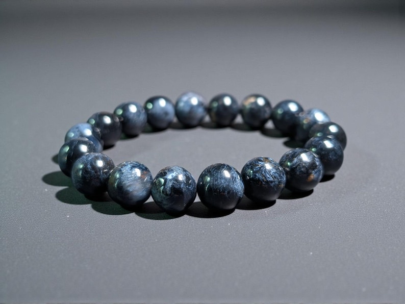 May include: A dark blue beaded bracelet. The beads are round and appear to be made of a polished stone. The bracelet is resting on a gray surface. The beads have a marbled appearance with lighter blue and white streaks.