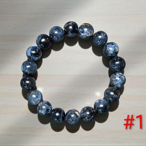 Puede incluir: Una pulsera circular compuesta por cuentas esf&eacute;ricas pulidas en tonos azul oscuro, gris y negro con una textura brillante y chatoyante. Las cuentas est&aacute;n ensartadas en un bucle continuo. El texto rojo #1 es visible en la parte inferior derecha.