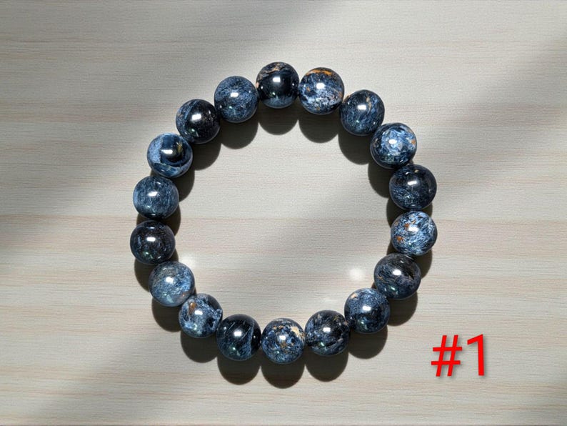 Puede incluir: Una pulsera circular compuesta por cuentas esf&eacute;ricas pulidas en tonos azul oscuro, gris y negro con una textura brillante y chatoyante. Las cuentas est&aacute;n ensartadas en un bucle continuo. El texto rojo #1 es visible en la parte inferior derecha.