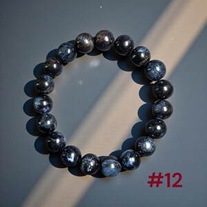 Puede incluir: Una pulsera circular de cuentas compuesta por piedras pulidas de color azul oscuro y negro con inclusiones fibrosas iridiscentes. Las cuentas est&aacute;n ensartadas en un bucle continuo. El texto #12 est&aacute; impreso en rojo en la esquina inferior derecha.