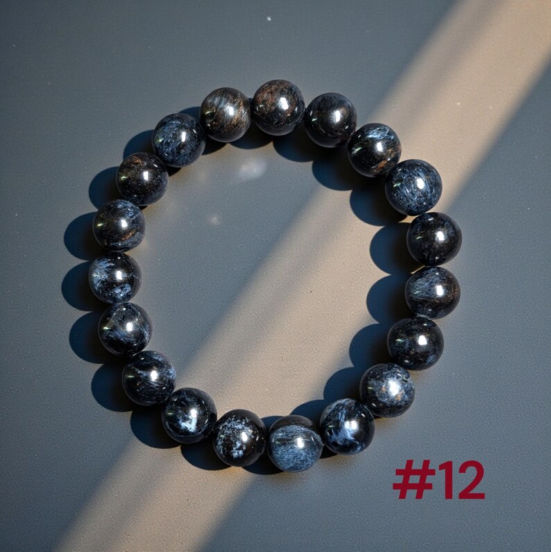 Puede incluir: Una pulsera circular de cuentas compuesta por piedras pulidas de color azul oscuro y negro con inclusiones fibrosas iridiscentes. Las cuentas est&aacute;n ensartadas en un bucle continuo. El texto #12 est&aacute; impreso en rojo en la esquina inferior derecha.