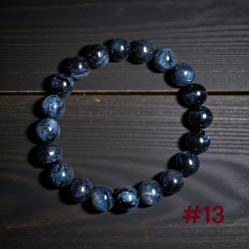Puede incluir: Una pulsera circular de cuentas compuesta por piedras esf&eacute;ricas pulidas de color azul y negro con un patr&oacute;n arremolinado y chatoyante. Las cuentas est&aacute;n ensartadas en un bucle continuo. El texto #13 es visible en la esquina inferior derecha.