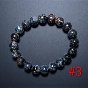 Puede incluir: Una pulsera circular compuesta por cuentas redondas y pulidas con un patr&oacute;n jaspeado en azul oscuro y gris con sutiles inclusiones met&aacute;licas en tonos cobre. Las cuentas est&aacute;n ensartadas en un bucle continuo. El texto rojo #3 es visible.