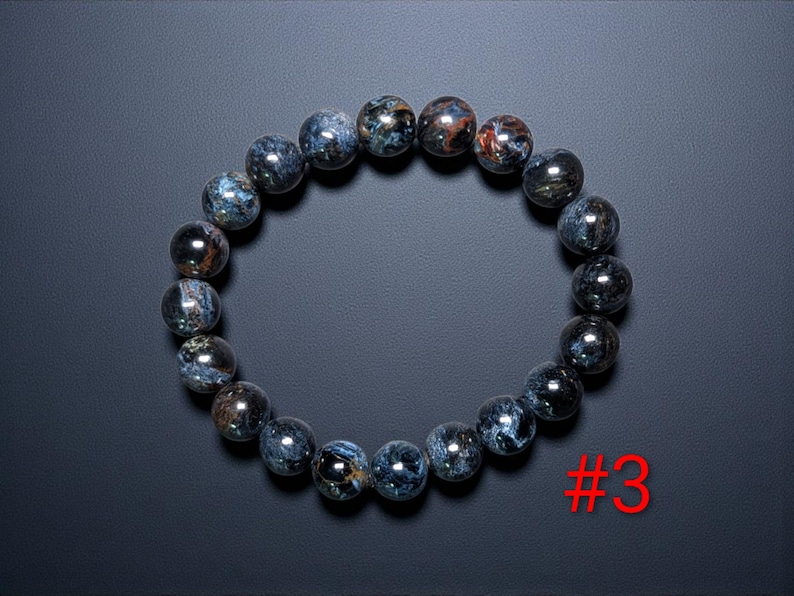 Puede incluir: Una pulsera circular compuesta por cuentas redondas y pulidas con un patr&oacute;n jaspeado en azul oscuro y gris con sutiles inclusiones met&aacute;licas en tonos cobre. Las cuentas est&aacute;n ensartadas en un bucle continuo. El texto rojo #3 es visible.
