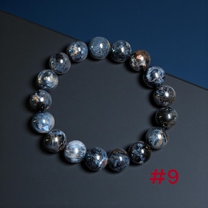 Puede incluir: Una pulsera circular compuesta por cuentas esf&eacute;ricas pulidas con un patr&oacute;n jaspeado en tonos azul profundo, gris y marr&oacute;n met&aacute;lico. Las cuentas est&aacute;n ensartadas en un bucle continuo y el n&uacute;mero 9 est&aacute; impreso en rojo en la esquina.