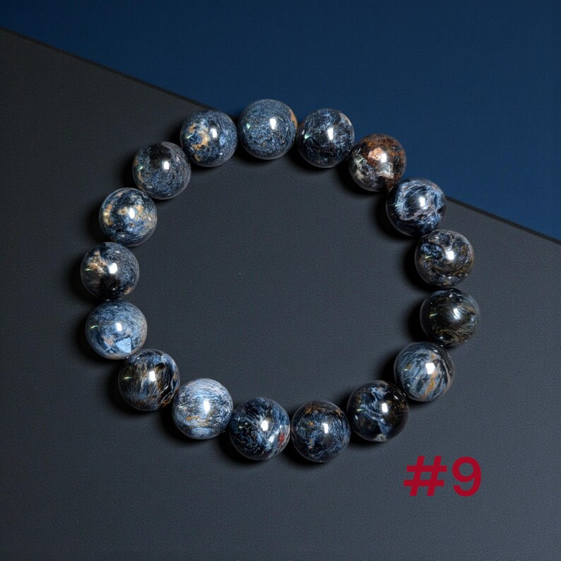 Puede incluir: Una pulsera circular compuesta por cuentas esf&eacute;ricas pulidas con un patr&oacute;n jaspeado en tonos azul profundo, gris y marr&oacute;n met&aacute;lico. Las cuentas est&aacute;n ensartadas en un bucle continuo y el n&uacute;mero 9 est&aacute; impreso en rojo en la esquina.