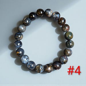 Puede incluir: Una pulsera el&aacute;stica circular compuesta por cuentas esf&eacute;ricas pulidas que presentan una mezcla de tonos azul profundo, gris carb&oacute;n y bronce met&aacute;lico con un brillo chatoyante. El n&uacute;mero #4 est&aacute; impreso en rojo en la esquina inferior derecha.