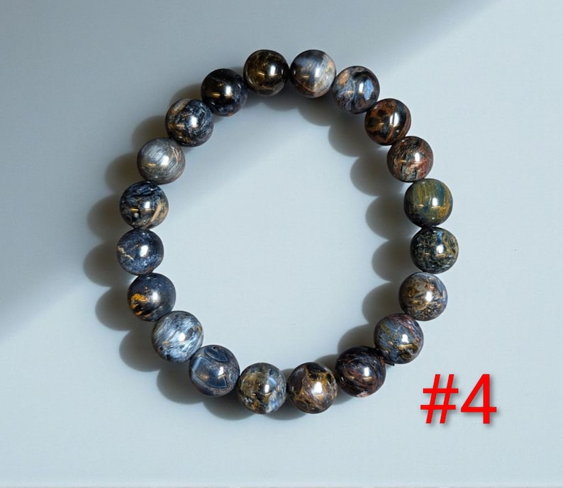 Puede incluir: Una pulsera el&aacute;stica circular compuesta por cuentas esf&eacute;ricas pulidas que presentan una mezcla de tonos azul profundo, gris carb&oacute;n y bronce met&aacute;lico con un brillo chatoyante. El n&uacute;mero #4 est&aacute; impreso en rojo en la esquina inferior derecha.