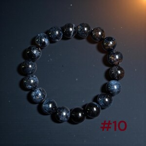 Puede incluir: Una pulsera circular de cuentas compuesta por piedras esf&eacute;ricas pulidas en tonos azul marino profundo, gris carb&oacute;n y negro con patrones fibrosos iridiscentes. El texto #10 est&aacute; impreso en rojo oscuro en la esquina inferior derecha.
