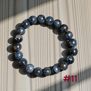 Puede incluir: Una pulsera circular compuesta por cuentas esf&eacute;ricas pulidas en tonos azul oscuro, gris y marr&oacute;n con un patr&oacute;n arremolinado y chatoyante. Las cuentas est&aacute;n ensartadas en un bucle continuo. El texto rojo #11 es visible en la parte inferior derecha.