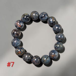 Puede incluir: Una pulsera circular compuesta por catorce cuentas esf&eacute;ricas pulidas con un patr&oacute;n de remolinos en tonos azul profundo, gris y cobre met&aacute;lico. Las cuentas est&aacute;n ensartadas en un bucle continuo y el n&uacute;mero 7 est&aacute; impreso en color rojo.