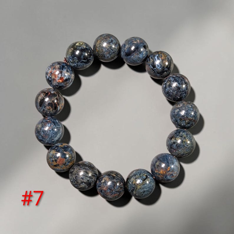Puede incluir: Una pulsera circular compuesta por catorce cuentas esf&eacute;ricas pulidas con un patr&oacute;n de remolinos en tonos azul profundo, gris y cobre met&aacute;lico. Las cuentas est&aacute;n ensartadas en un bucle continuo y el n&uacute;mero 7 est&aacute; impreso en color rojo.