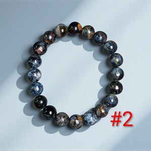 Puede incluir: Una pulsera circular de cuentas compuesta por piedras esf&eacute;ricas pulidas con un patr&oacute;n jaspeado de tonos azul profundo, negro y marr&oacute;n dorado. La pulsera est&aacute; dispuesta en forma de anillo e incluye el texto rojo #2 en la esquina inferior derecha.
