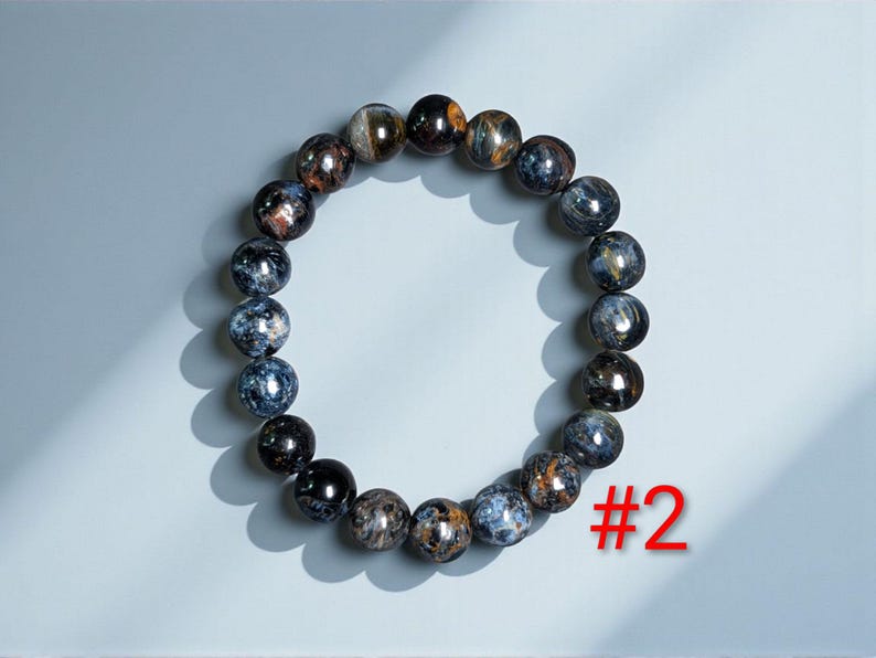 Puede incluir: Una pulsera circular de cuentas compuesta por piedras esf&eacute;ricas pulidas con un patr&oacute;n jaspeado de tonos azul profundo, negro y marr&oacute;n dorado. La pulsera est&aacute; dispuesta en forma de anillo e incluye el texto rojo #2 en la esquina inferior derecha.